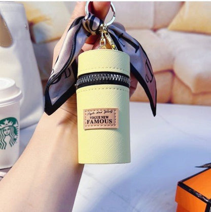 Pastel yellow keychain pouch with 'Vogue New Famous' label, silk ribbon, and zipper, held in hand near coffee cup